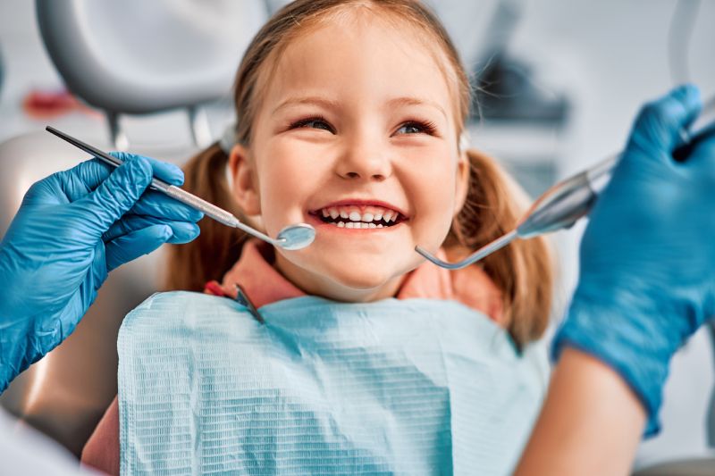Smiling young girl with dental bib at dentist showing teeth during checkup with dental tools.