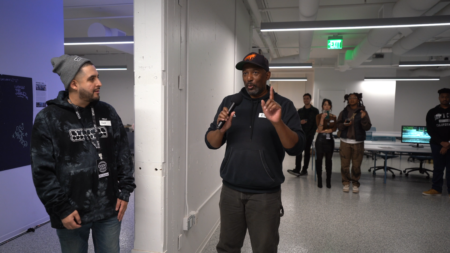 A speaker in a cap talks beside another presenter.