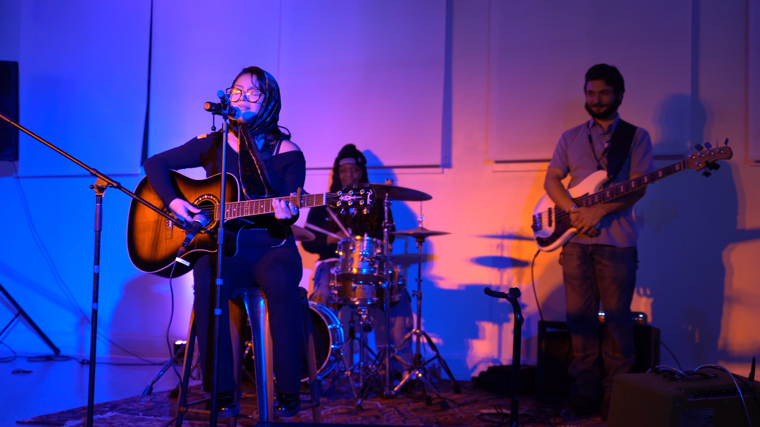 A singer plays acoustic guitar with a drummer and bassist.