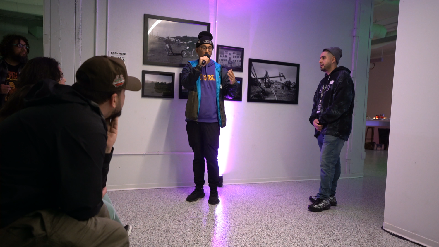Two performers stand in a purple-lit exhibition space.