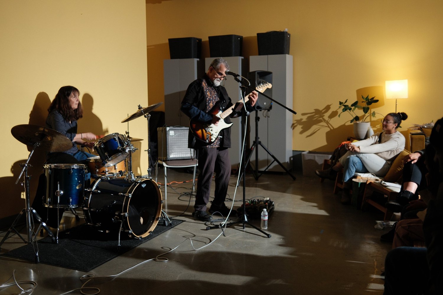 Guitarist and drummer playing for a closely seated audience.