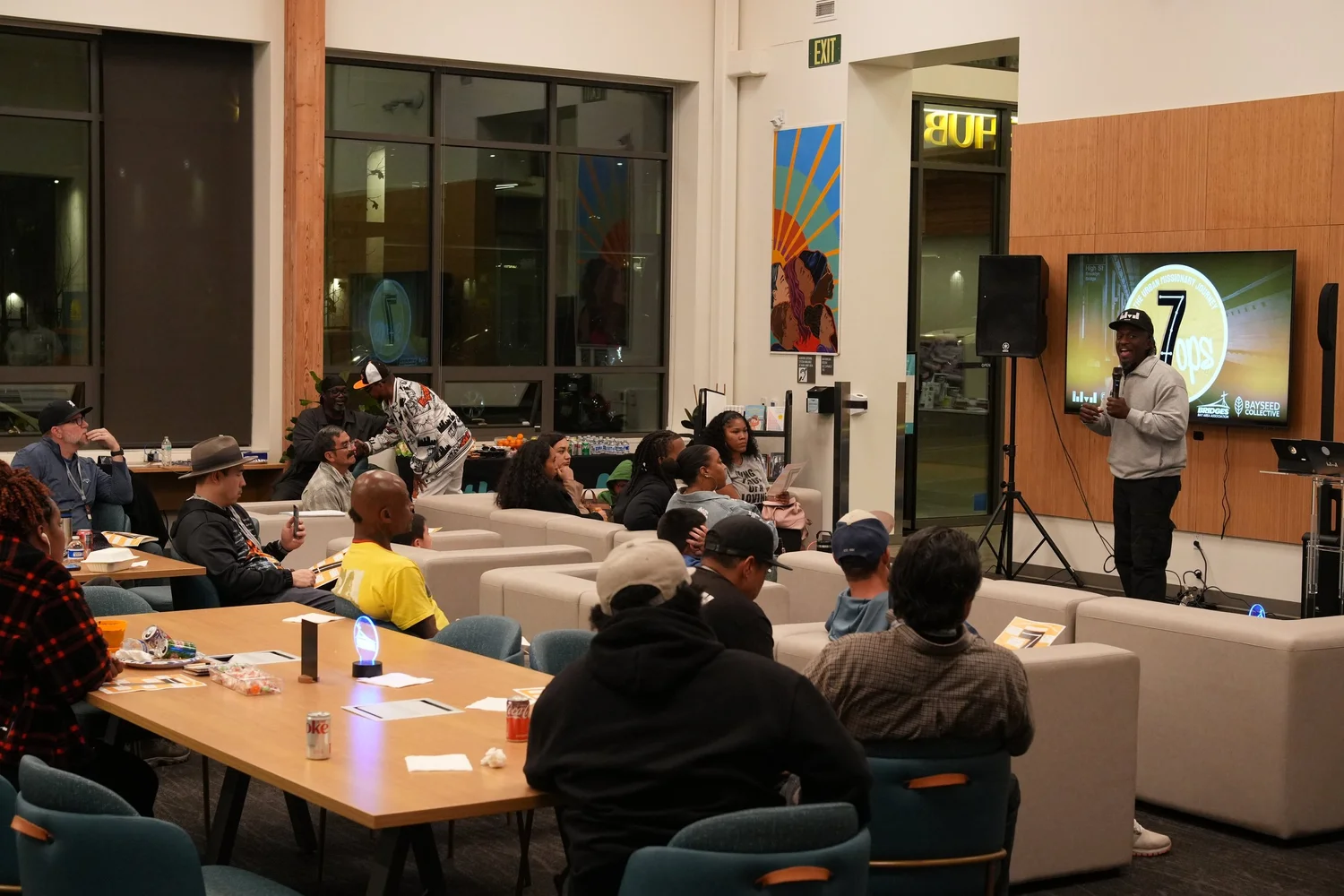 Guests seated around tables and sofas listen to the community program.
