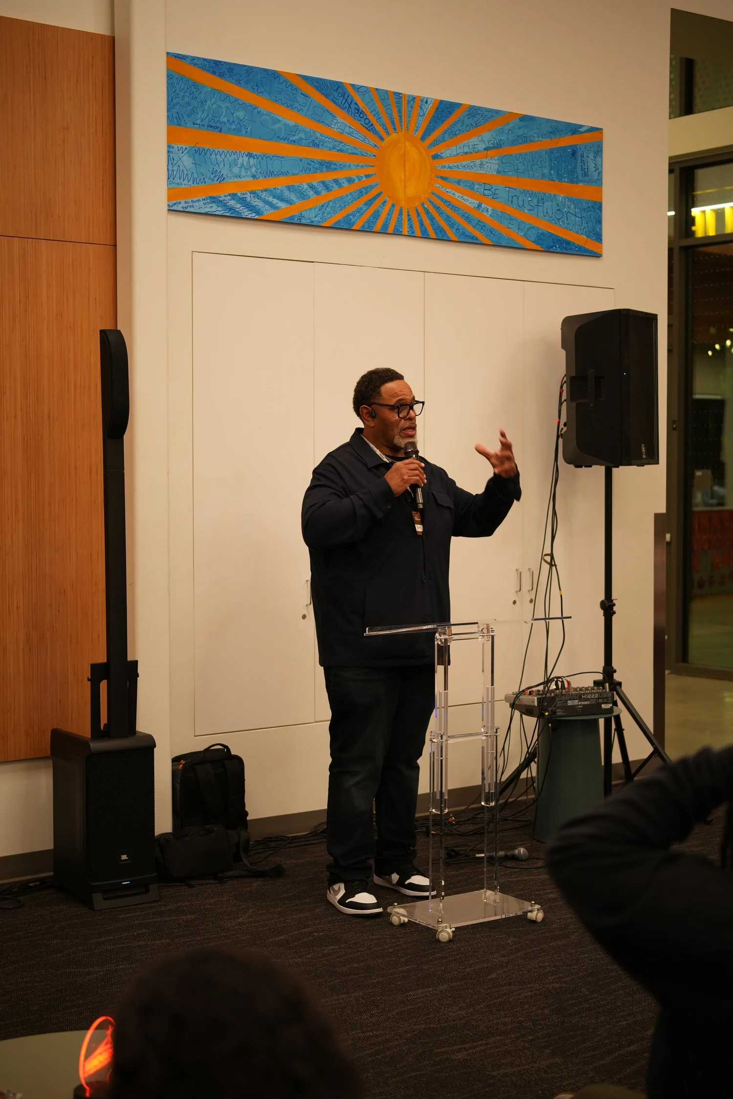 Speaker gesturing with both hands near a wall-mounted speaker.
