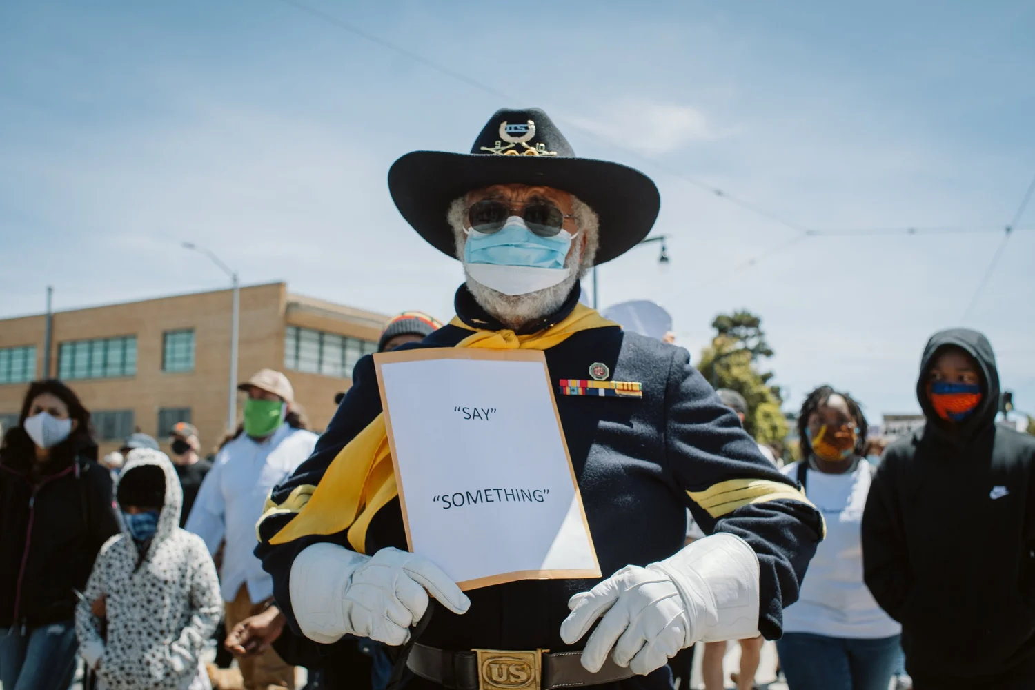 Masked participant in a wide-brimmed hat holding a Say Something sign.