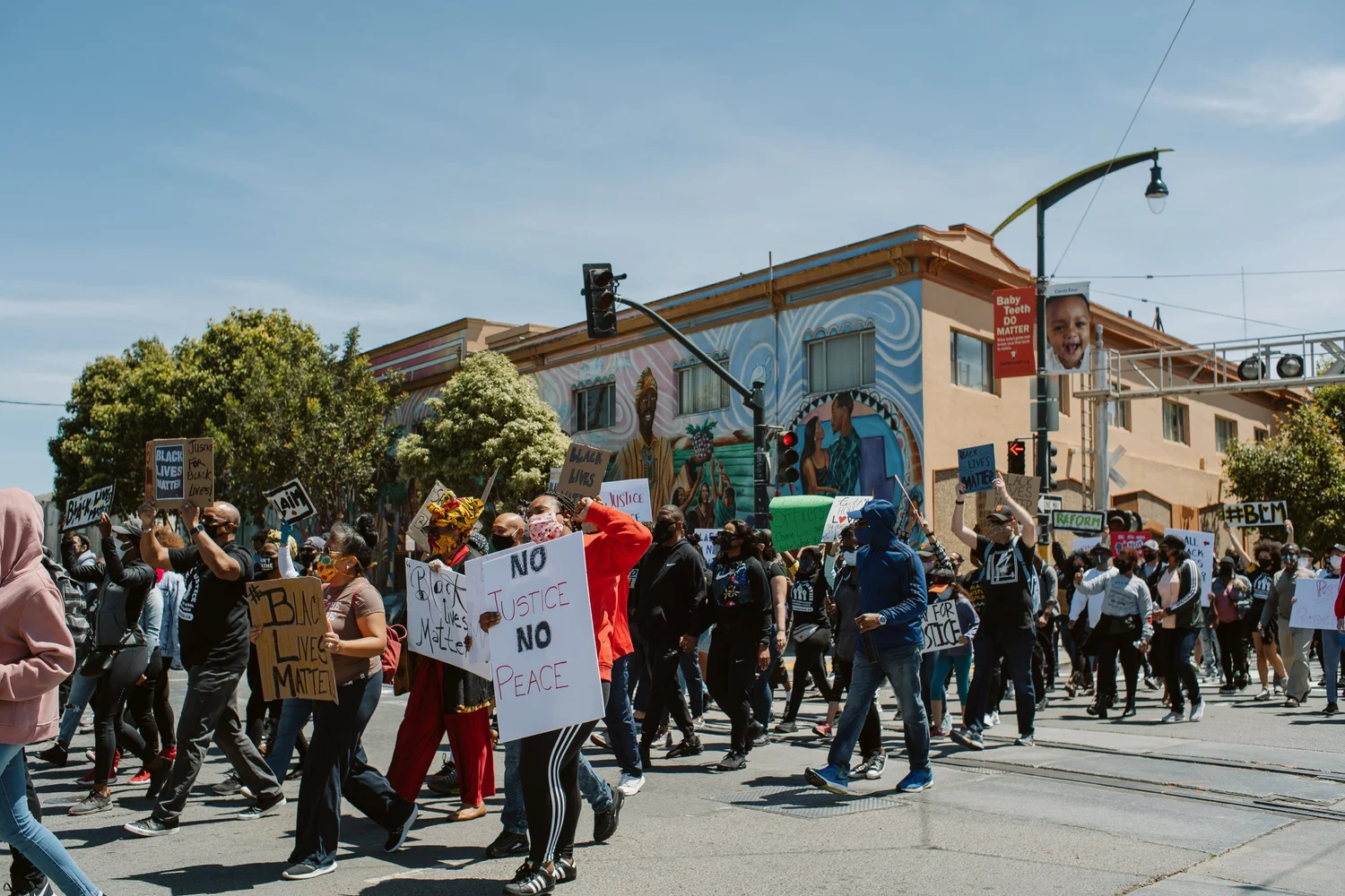 Large procession passing a mural-covered corner building.