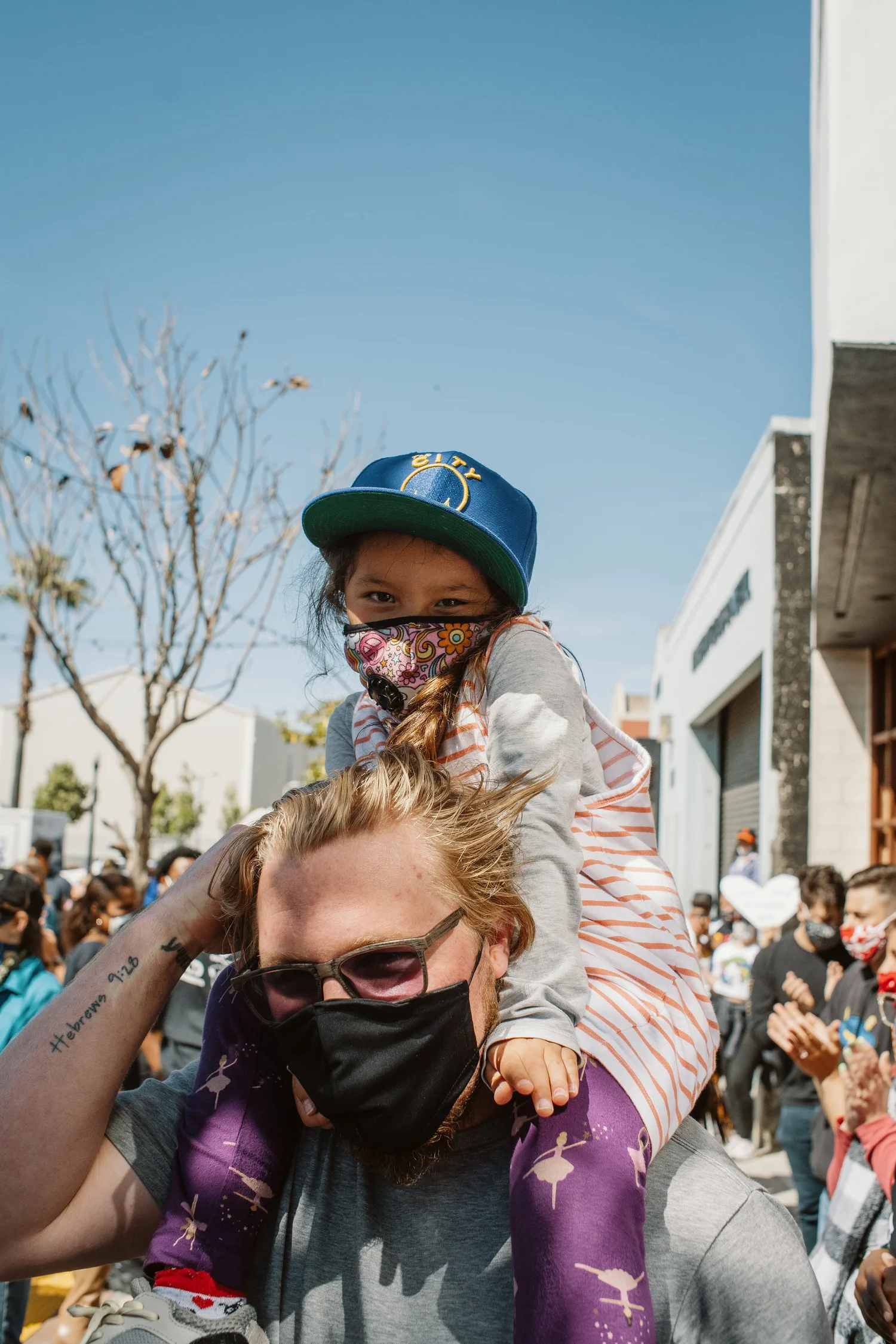 Child wearing a cap sitting on an adult's shoulders.