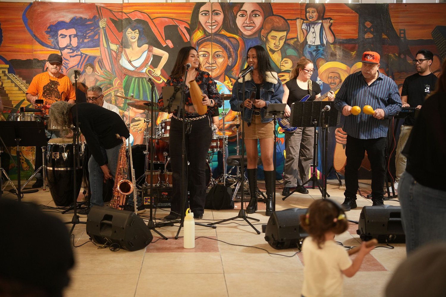 Vocalists and instrumentalists perform before an audience and a colorful mural.