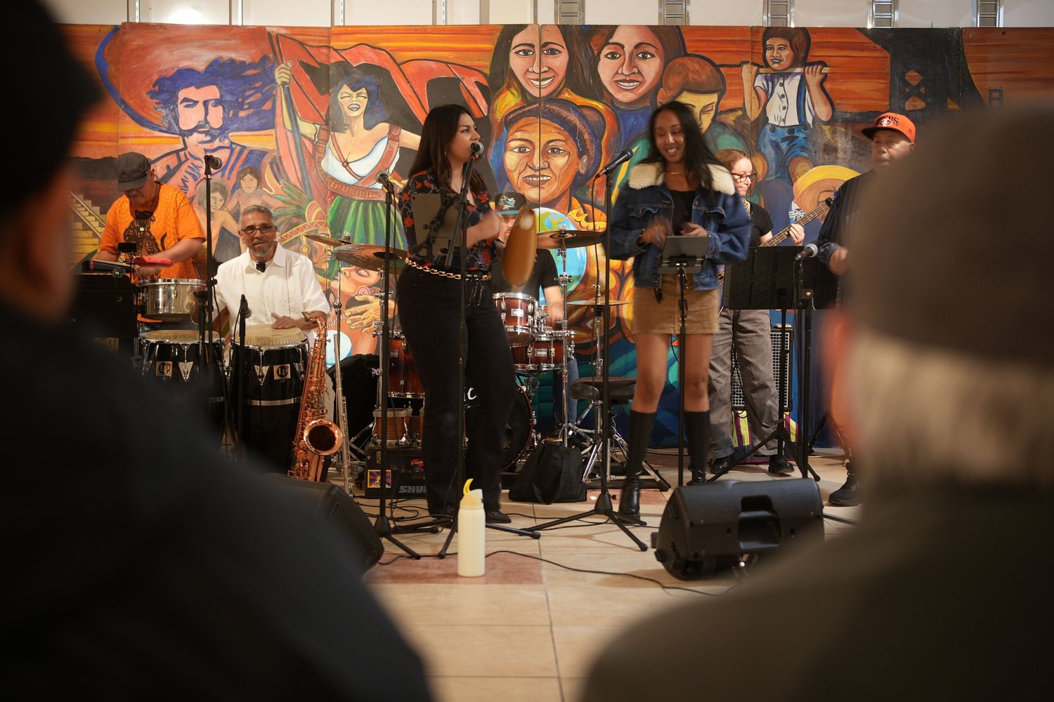 Vocalists and instrumentalists perform before an audience and a colorful mural.