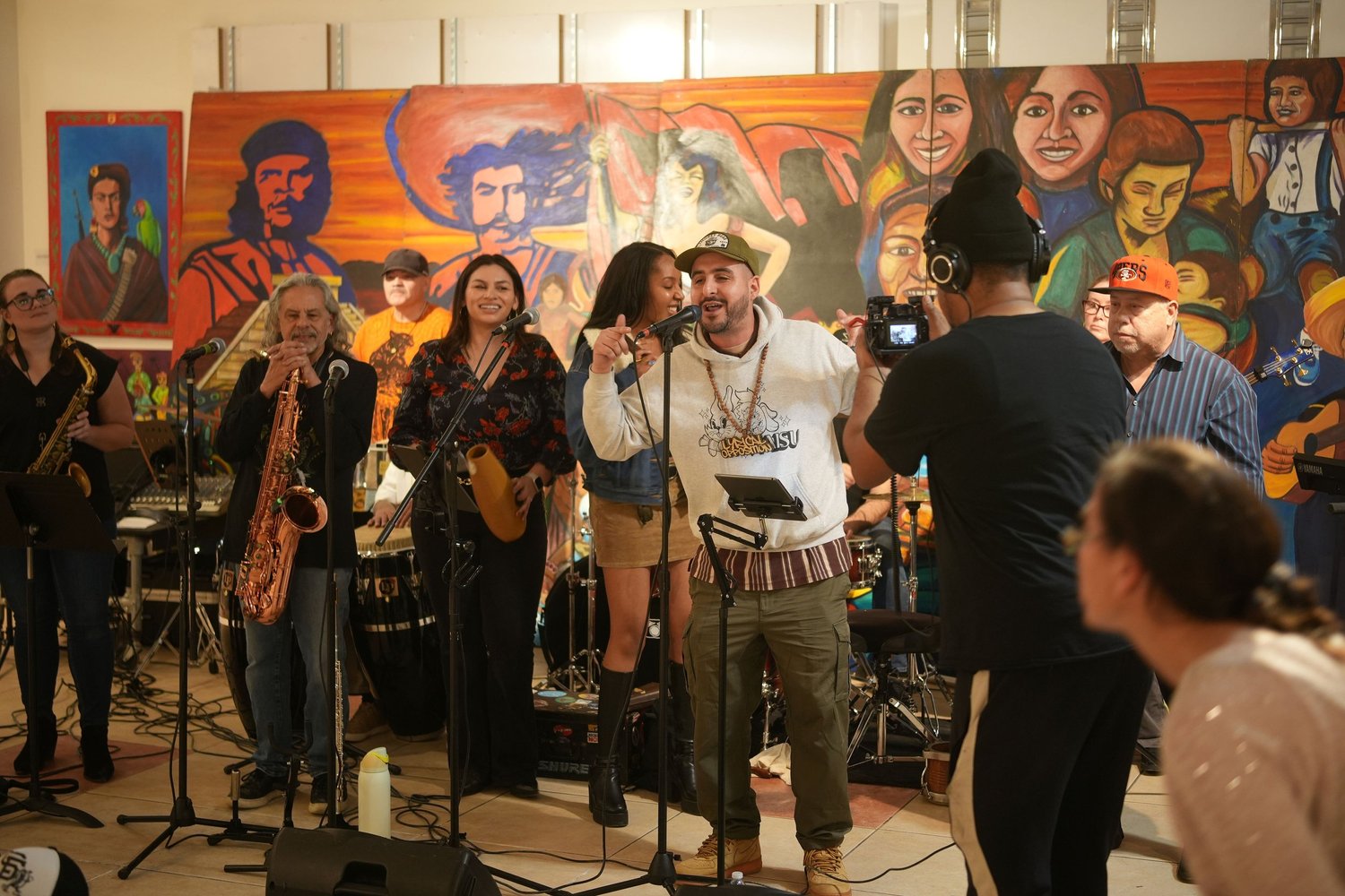 Vocalists and instrumentalists perform before an audience and a colorful mural.