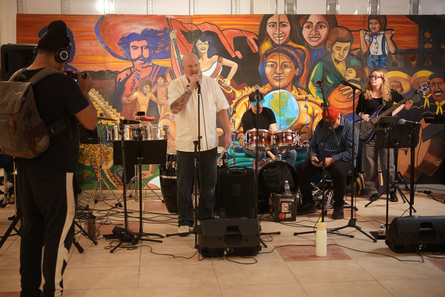 Vocalists and instrumentalists perform before an audience and a colorful mural.