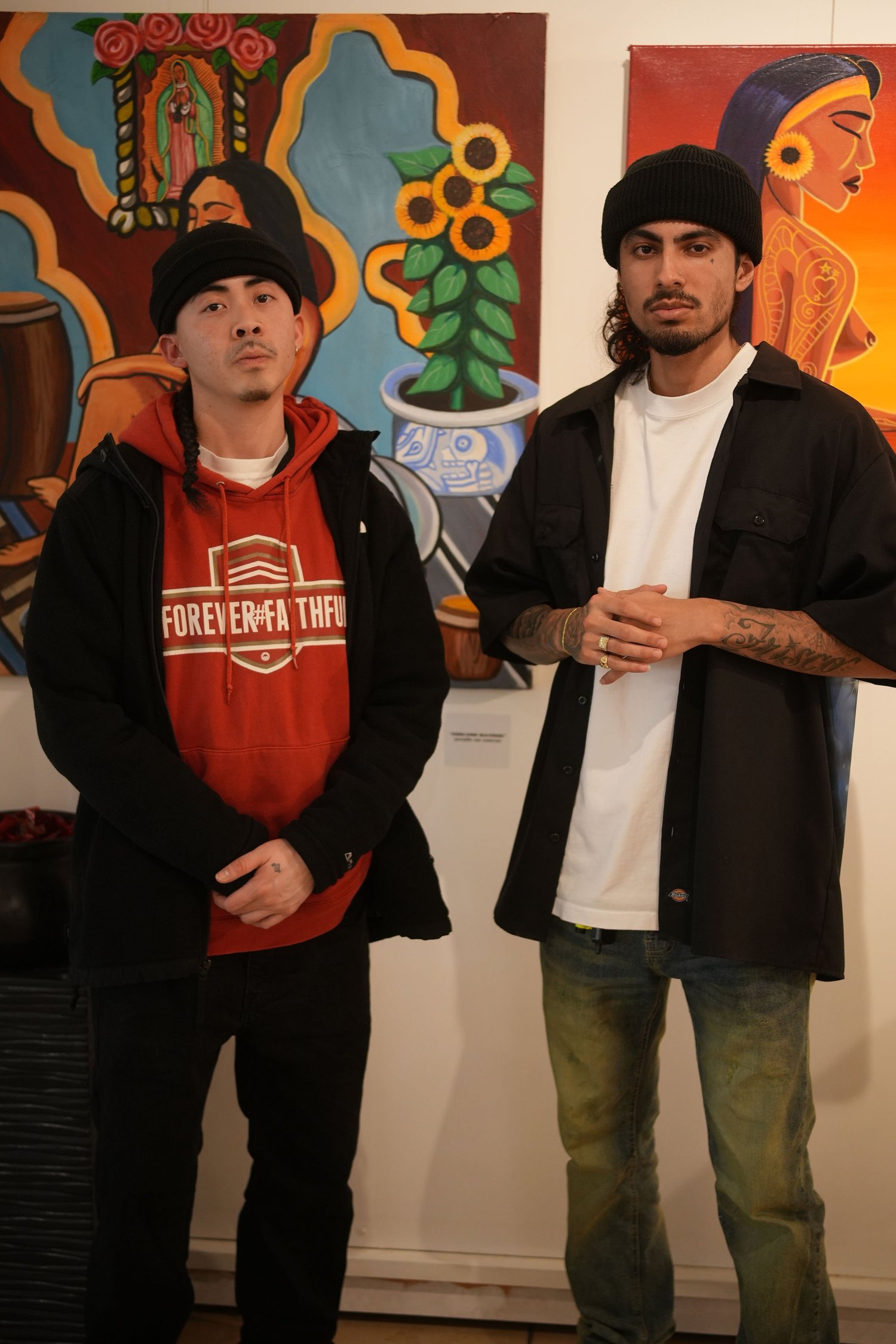 Two artists pose in front of brightly colored paintings.