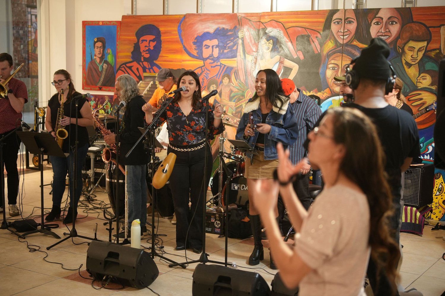 Vocalists and instrumentalists perform before an audience and a colorful mural.
