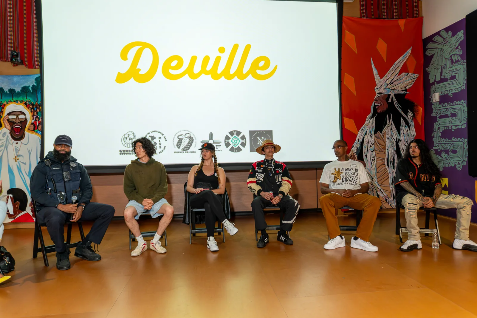 Panel participants sit beneath the projected Deville title, with murals and blue curtains around them.