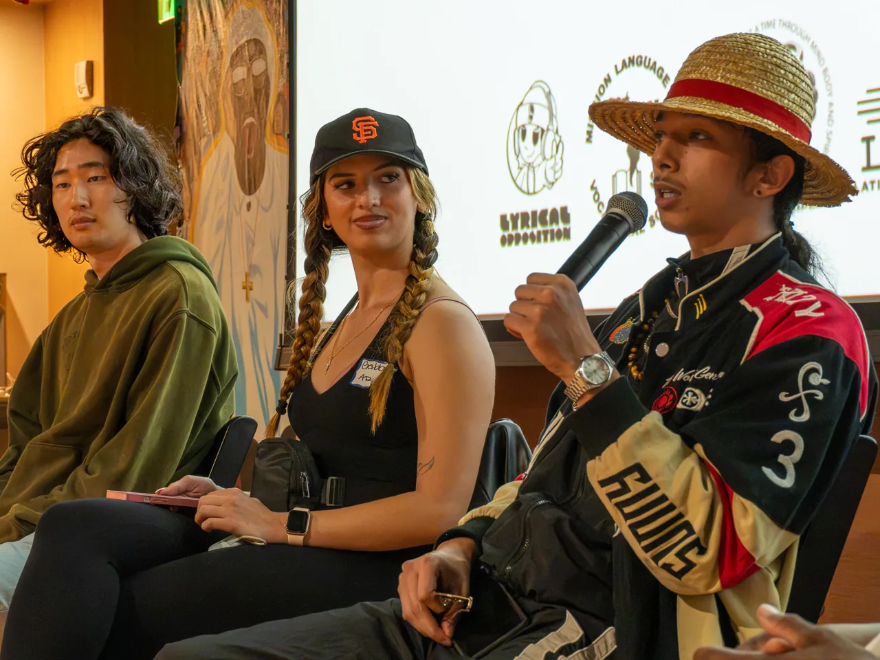 A panel participant in a straw hat speaks while two seated participants listen.