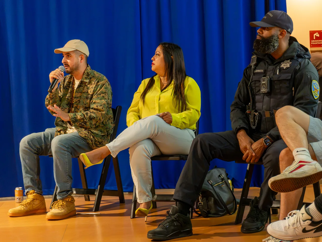 A panel participant in a patterned jacket and cap speaks beside participants in yellow and black.