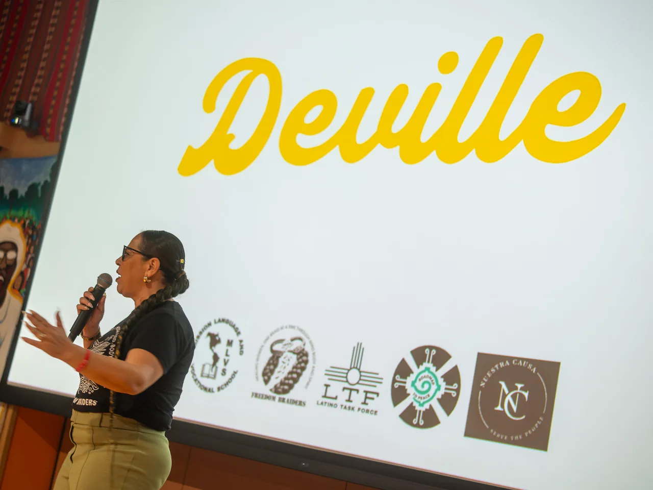 A speaker in a black shirt and olive trousers addresses guests beneath the Deville title.