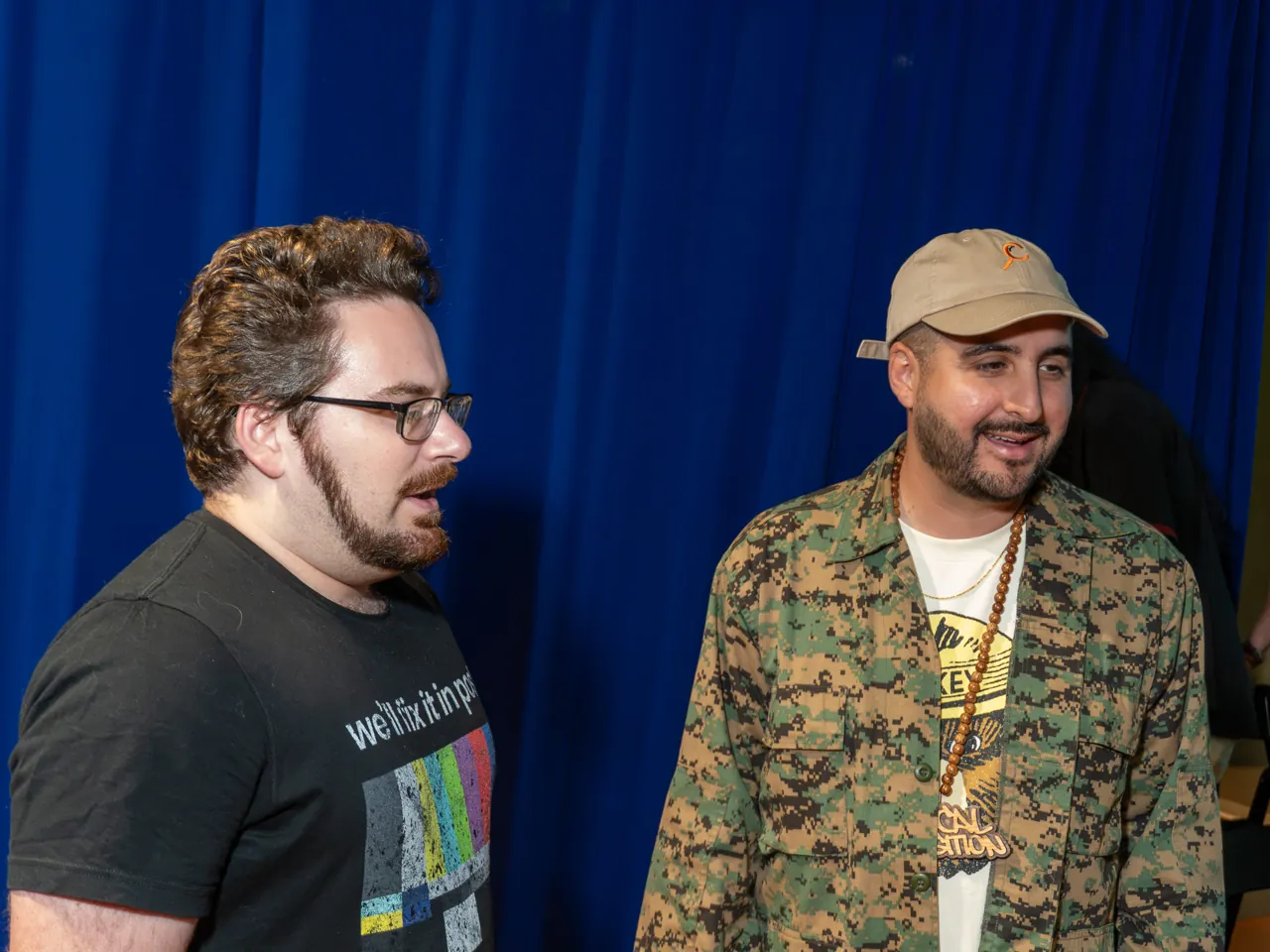 Two guests talk beside the blue curtain, one wearing a patterned jacket and cap.