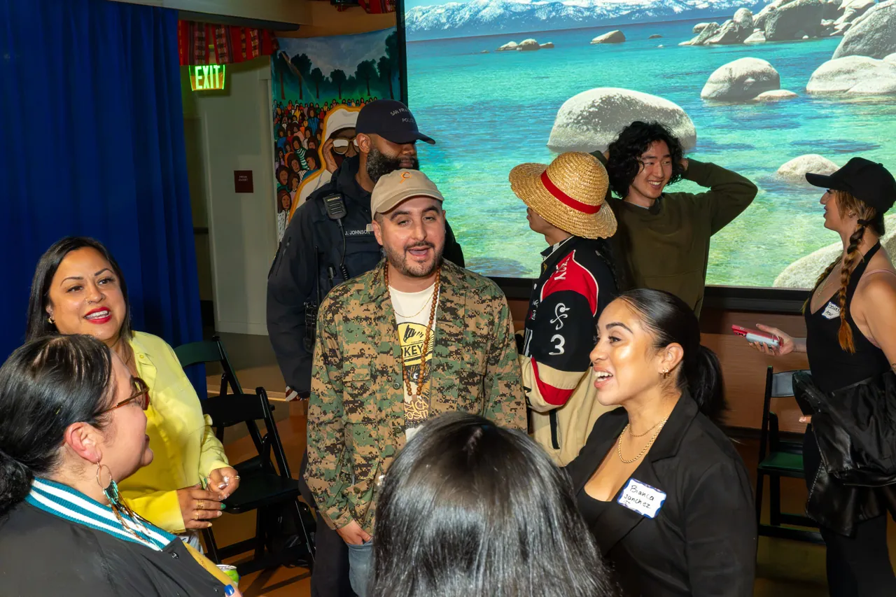 Guests gather closely around a person in a patterned jacket and cap to talk.