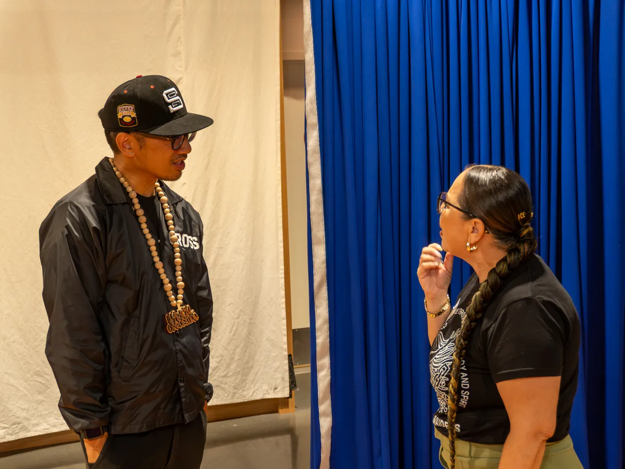 Two guests talk beside the blue curtain, one wearing a black cap and jacket.