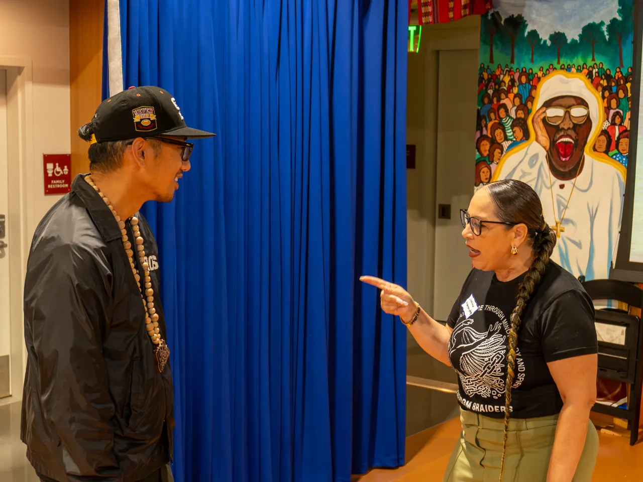 Two guests talk beside the blue curtain, one wearing a black cap and jacket.