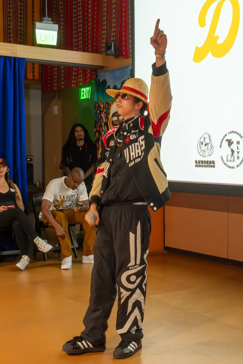 A performer raises one arm while holding a microphone beside the Deville projection.