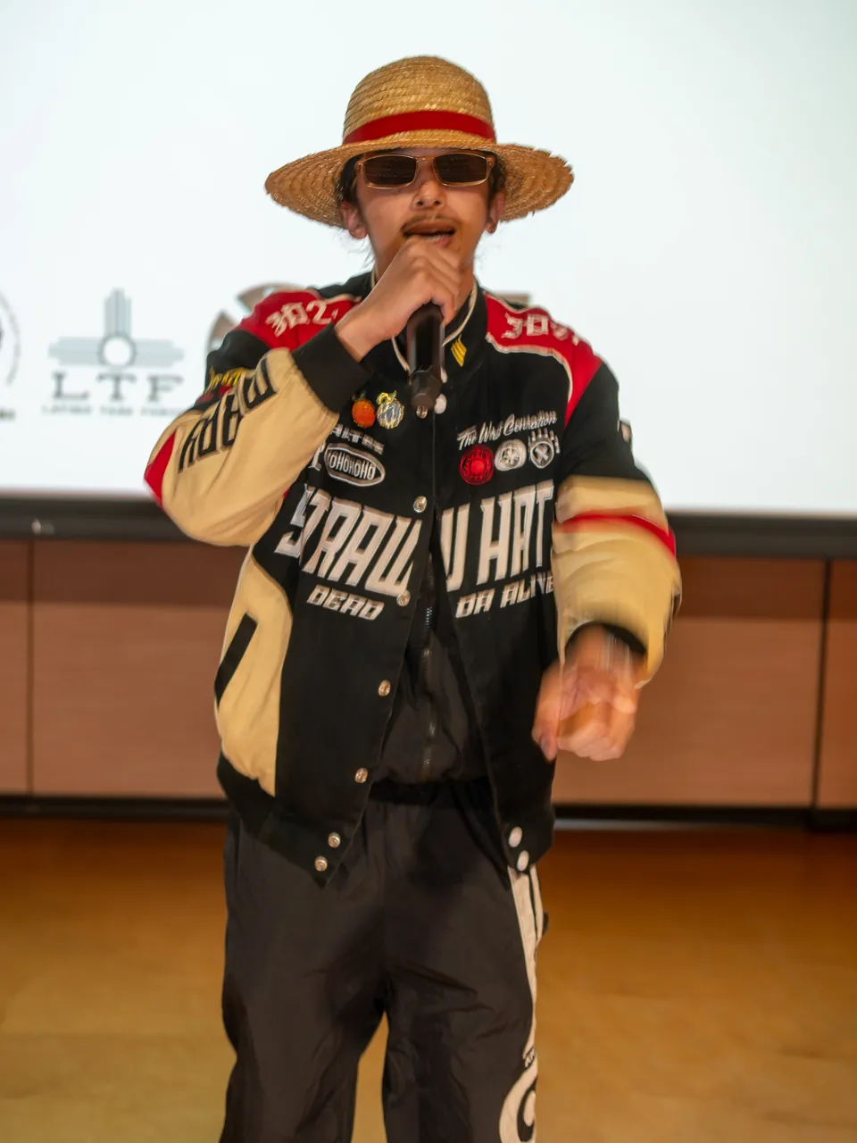 A performer in a straw hat and racing jacket holds a microphone in front of the Deville screen.