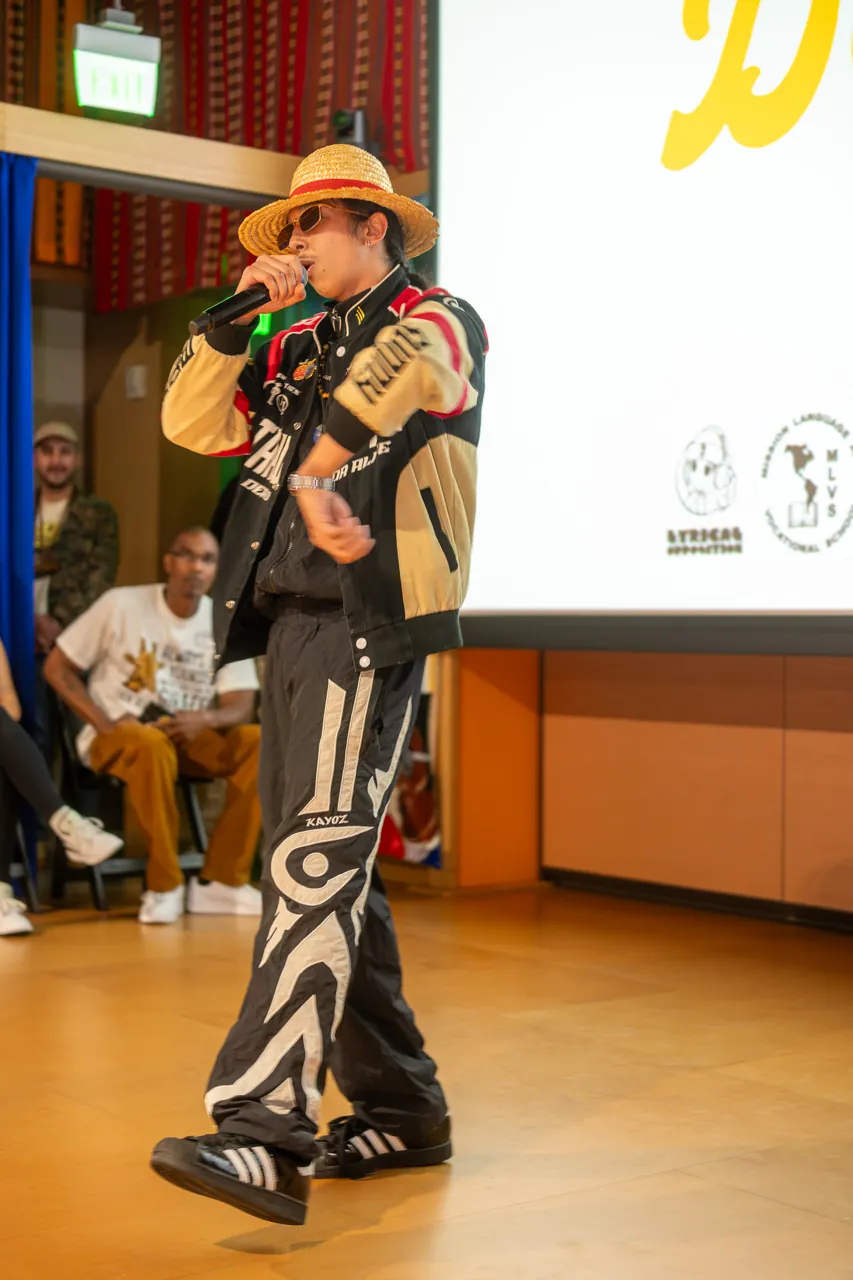 A performer in a straw hat and racing jacket holds a microphone in front of the Deville screen.