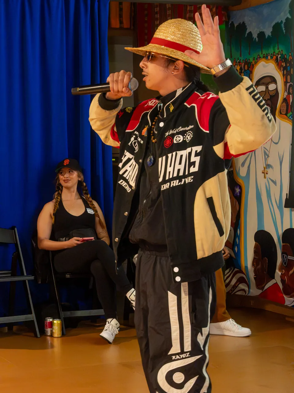 A performer in a straw hat and racing jacket holds a microphone in front of the Deville screen.