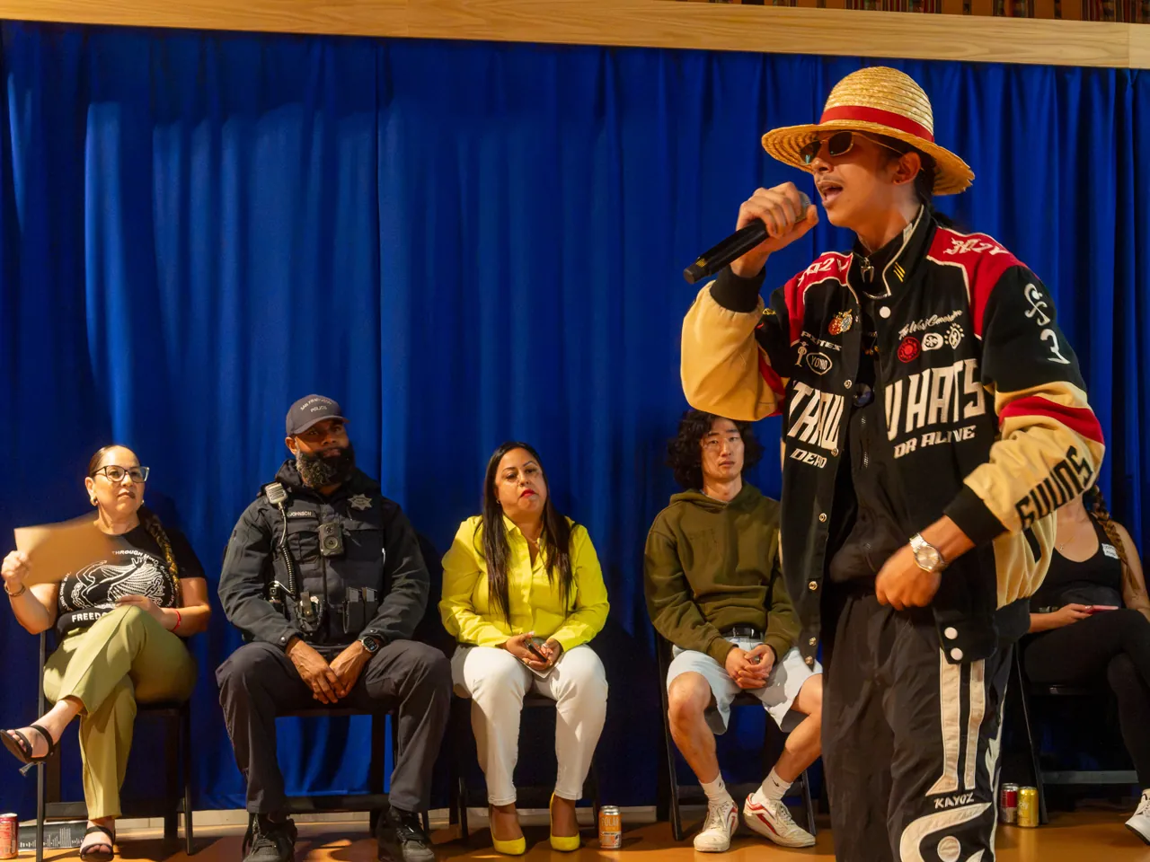 A performer in a straw hat and racing jacket holds a microphone in front of the Deville screen.