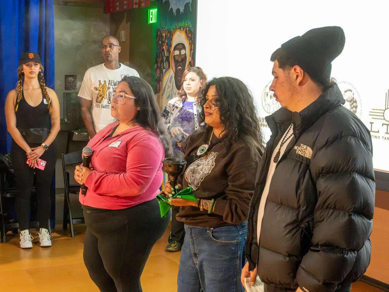 Several people stand together at the front of the room, including a person in pink and a person holding green fabric.