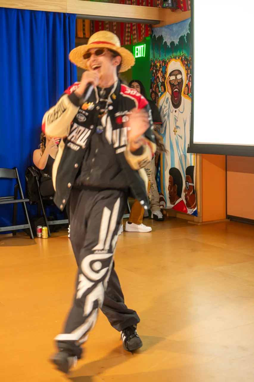 A performer in a straw hat and racing jacket holds a microphone in front of the Deville screen.