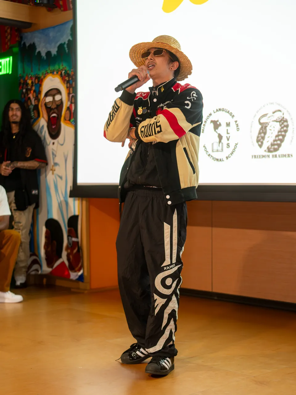 A performer in a straw hat and racing jacket holds a microphone in front of the Deville screen.