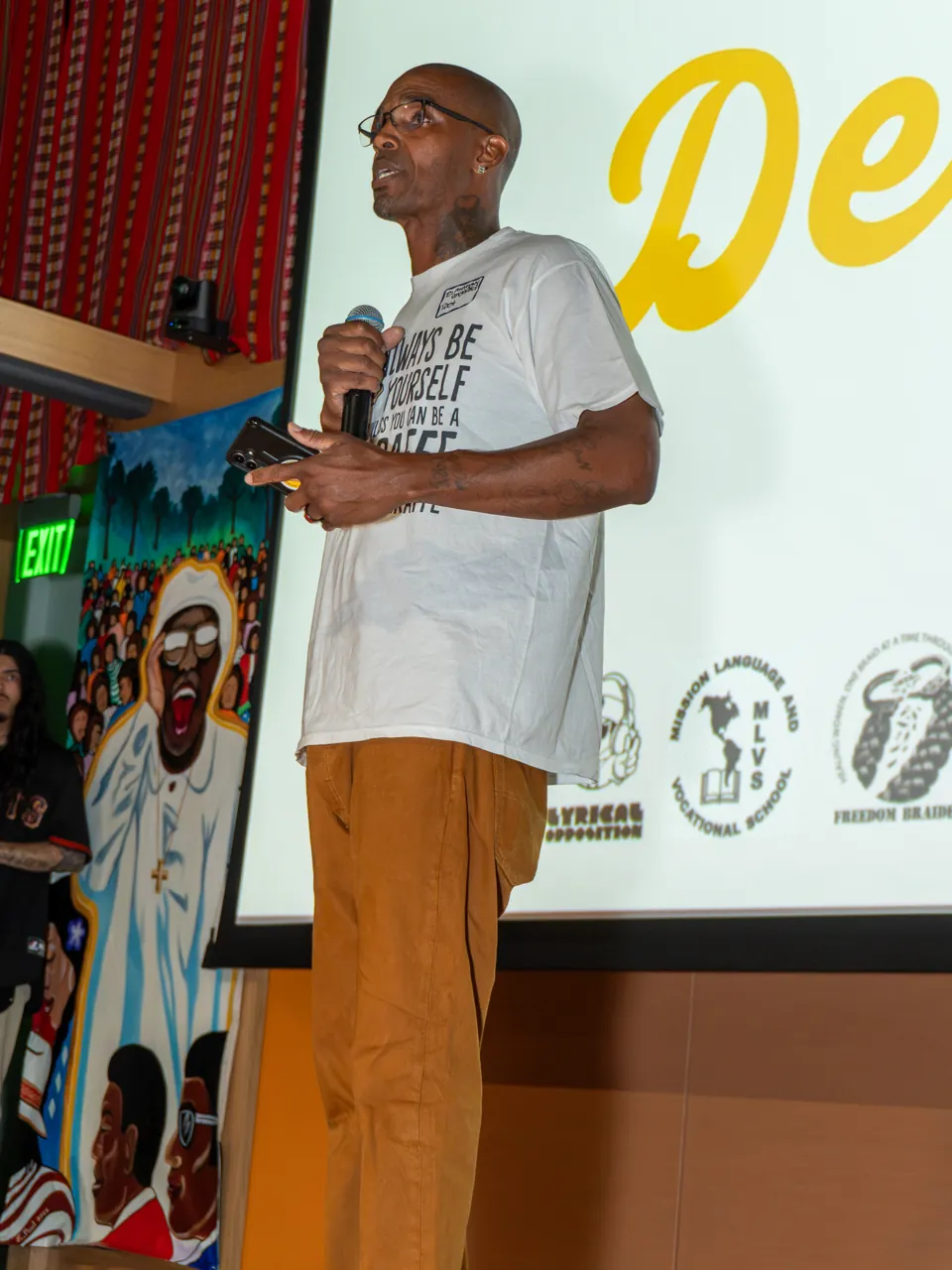 A speaker in a white T-shirt looks down at a phone while holding a microphone.