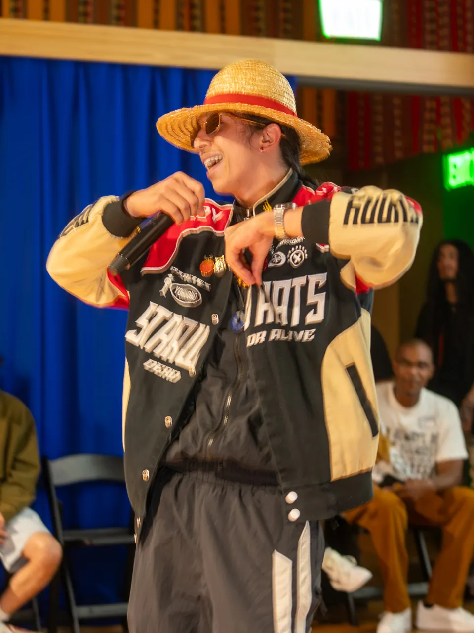 A performer in a straw hat and racing jacket holds a microphone in front of the Deville screen.