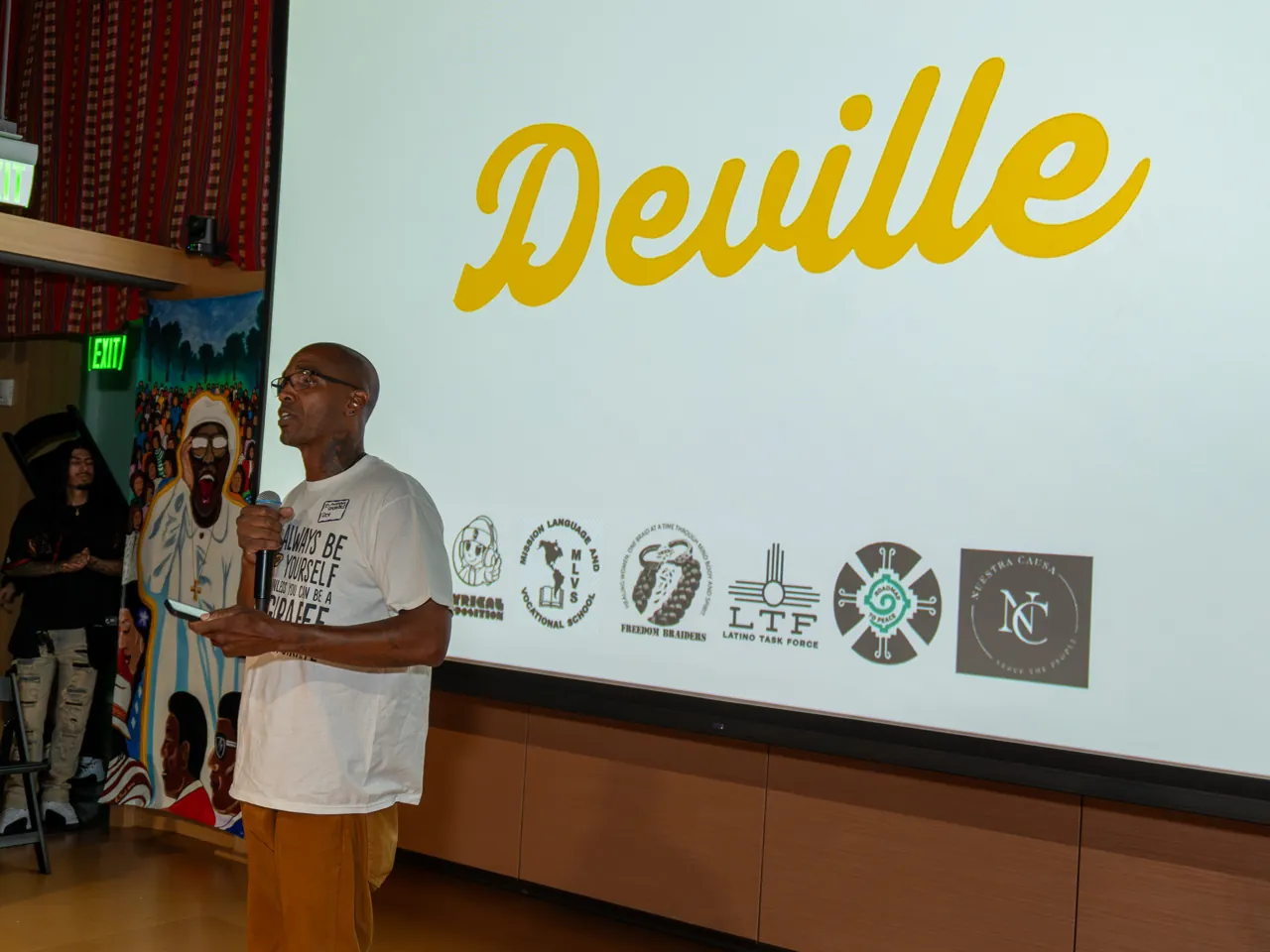 A speaker in a white T-shirt and mustard trousers addresses the room with a microphone.