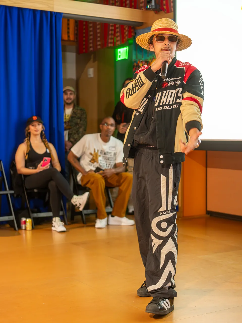 A performer in a straw hat and racing jacket holds a microphone in front of the Deville screen.