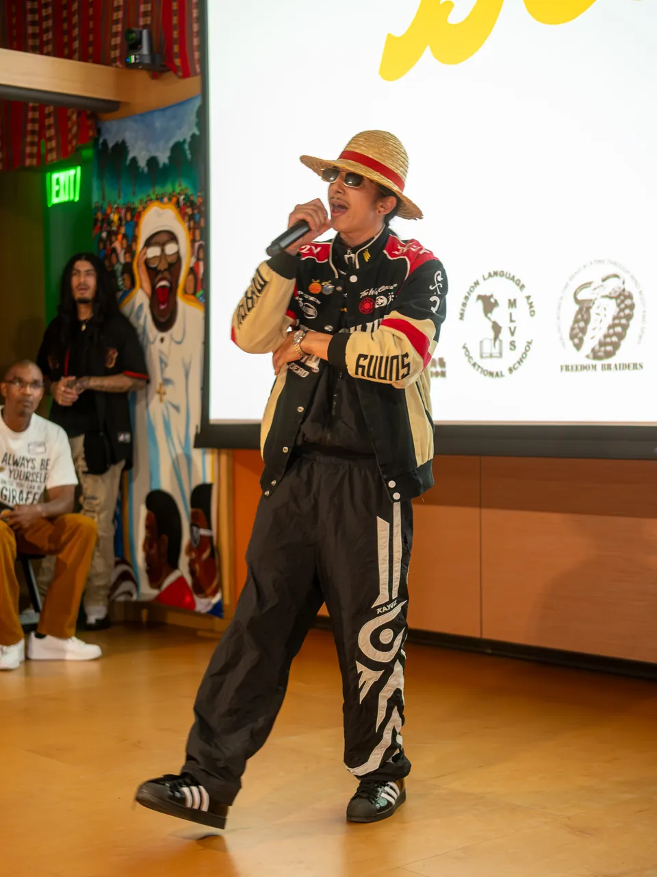 A performer in a straw hat and racing jacket holds a microphone in front of the Deville screen.