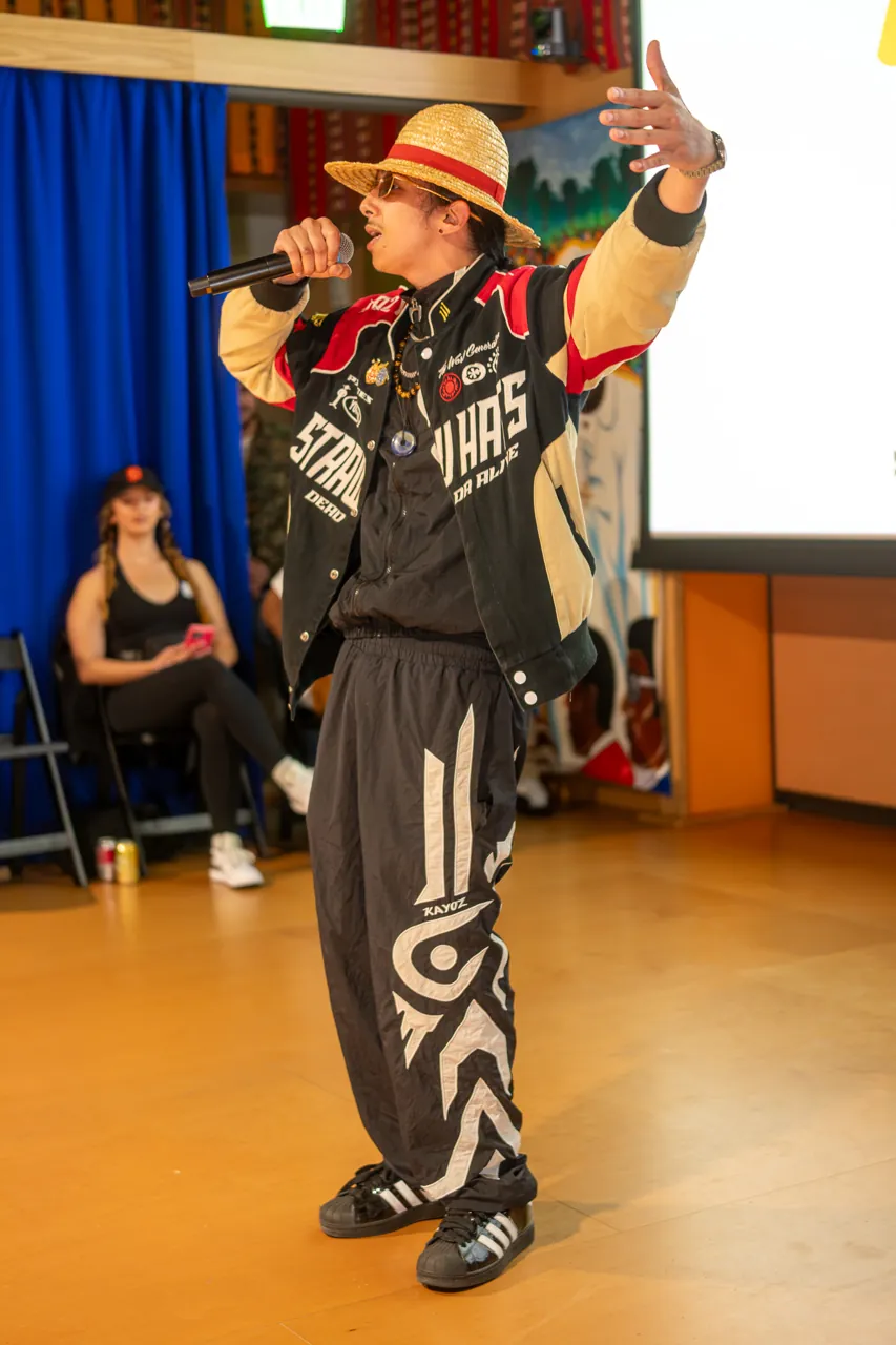 A performer raises one arm while holding a microphone beside the Deville projection.