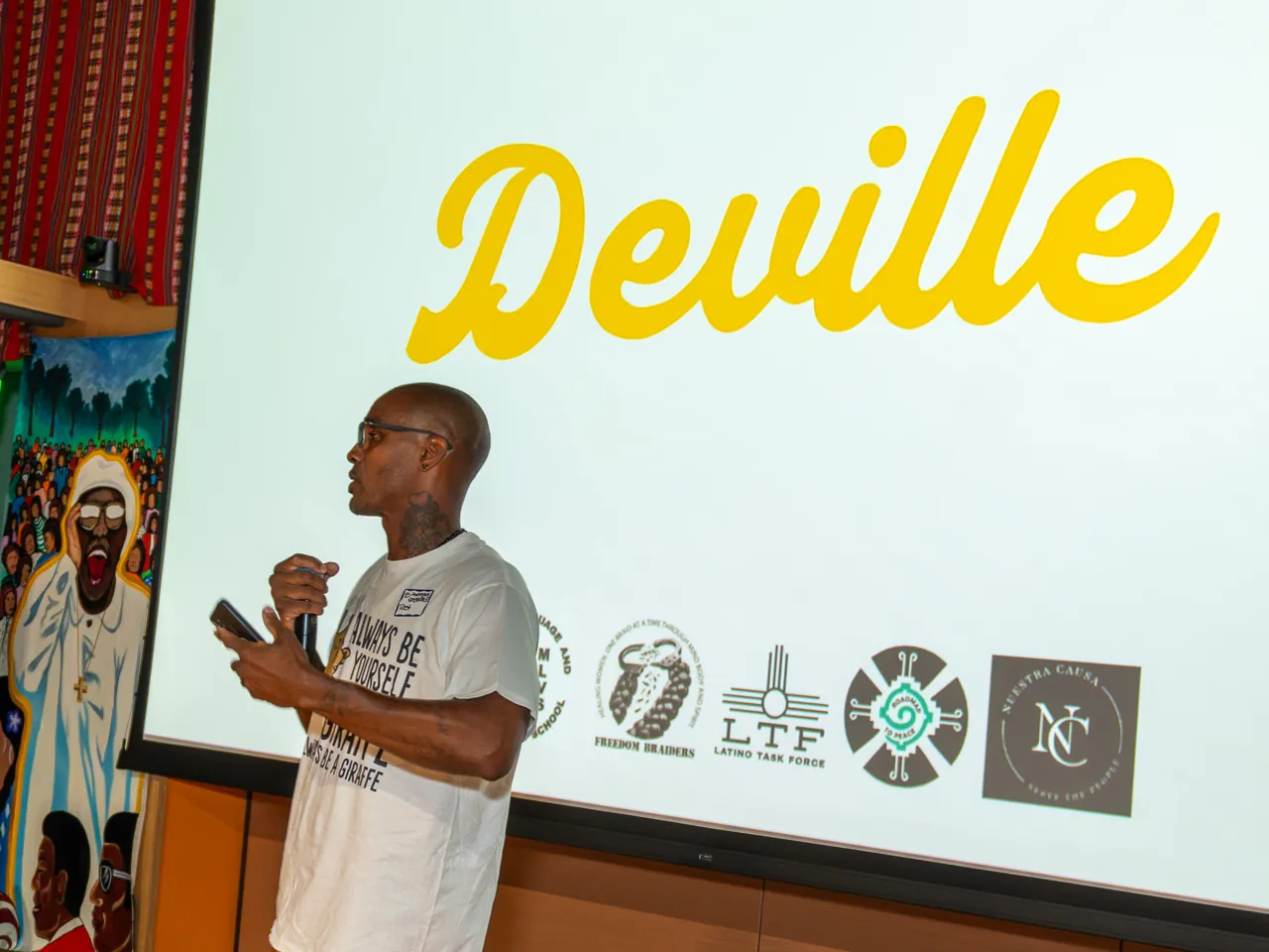 A speaker in a white T-shirt and mustard trousers addresses the room with a microphone.