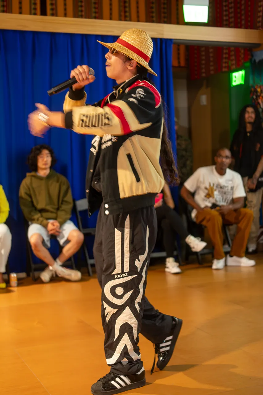 A performer in a straw hat reaches toward the audience while speaking into a microphone.