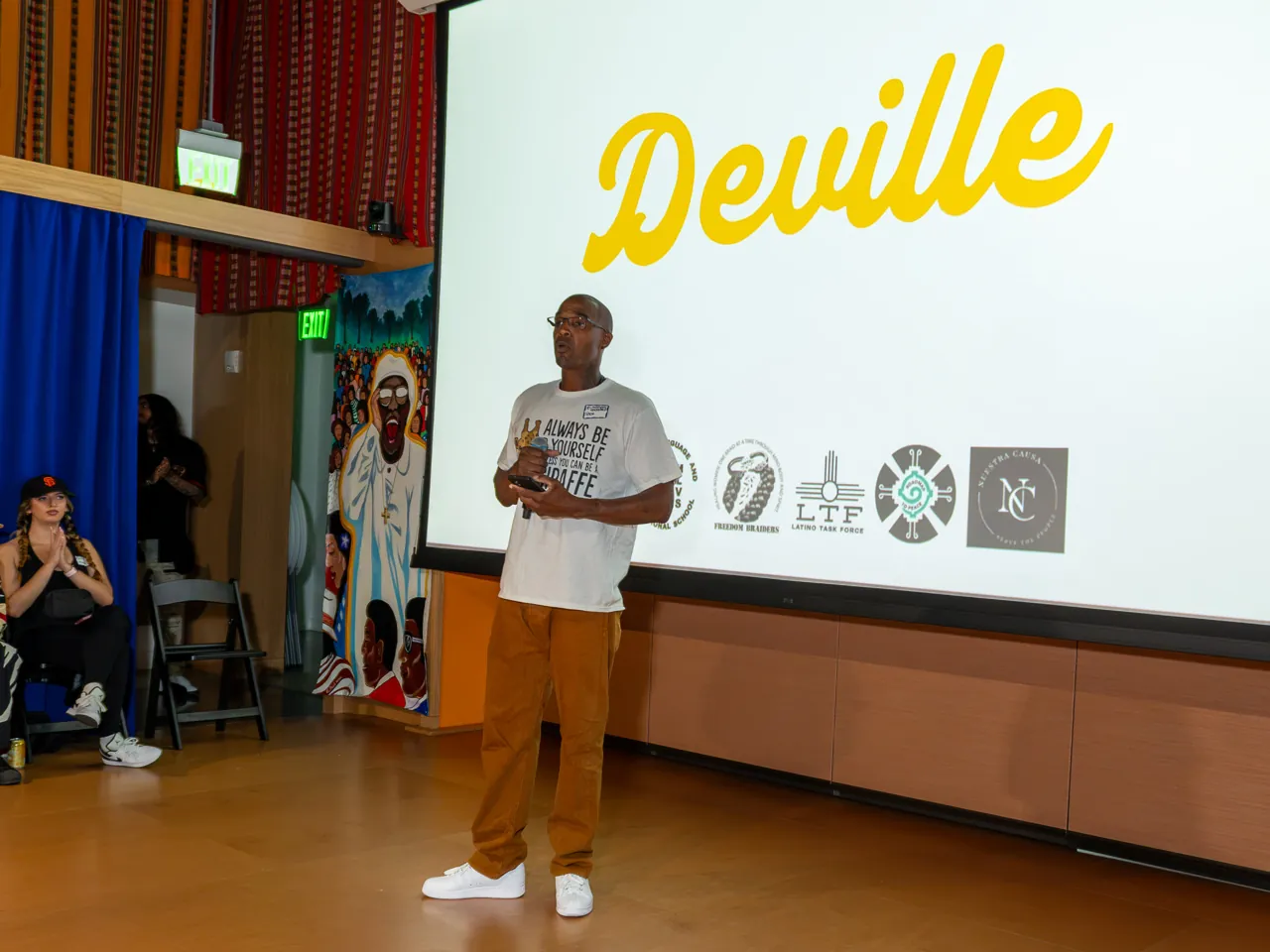 A speaker in a white T-shirt and mustard trousers addresses the room with a microphone.