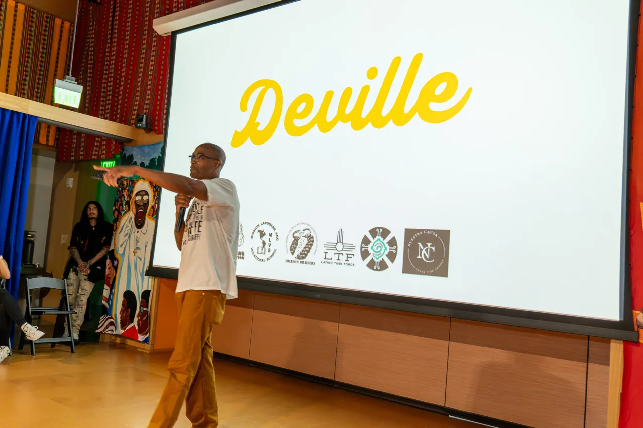 A speaker in a white T-shirt extends an arm toward the audience beneath the Deville title.