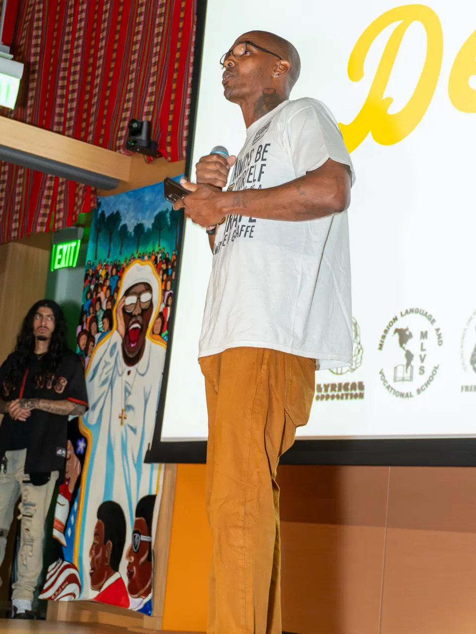 A speaker in a white T-shirt gestures toward the audience with an outstretched arm.