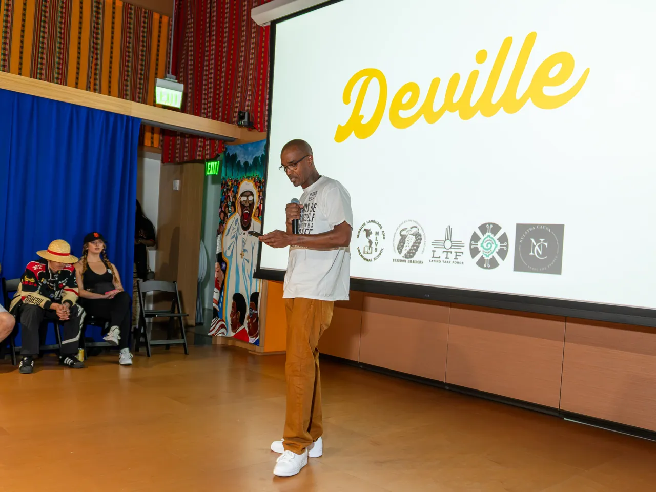 A speaker in a white T-shirt and mustard trousers addresses the room with a microphone.