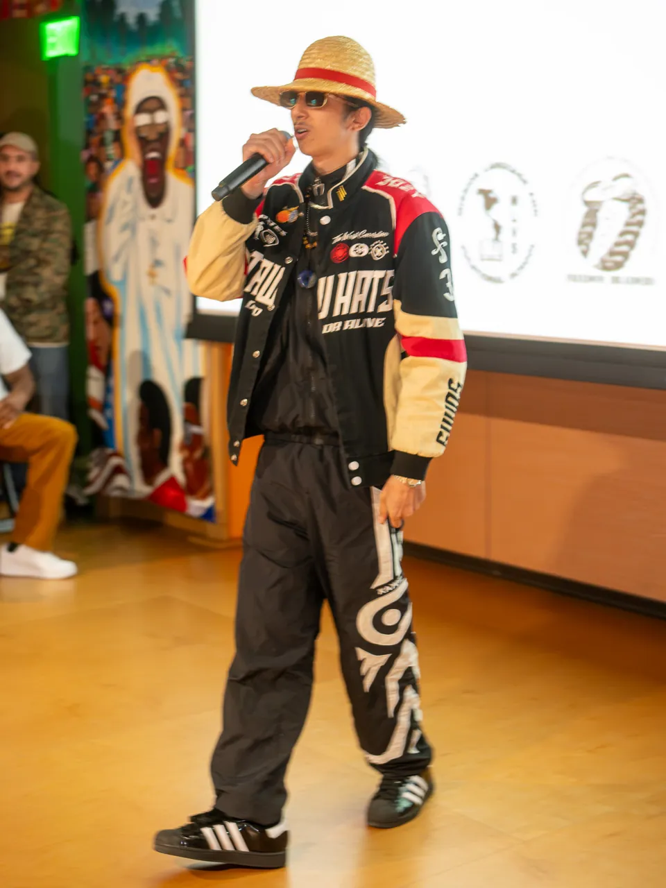 A performer in a straw hat and racing jacket delivers into a microphone beside the projection screen.
