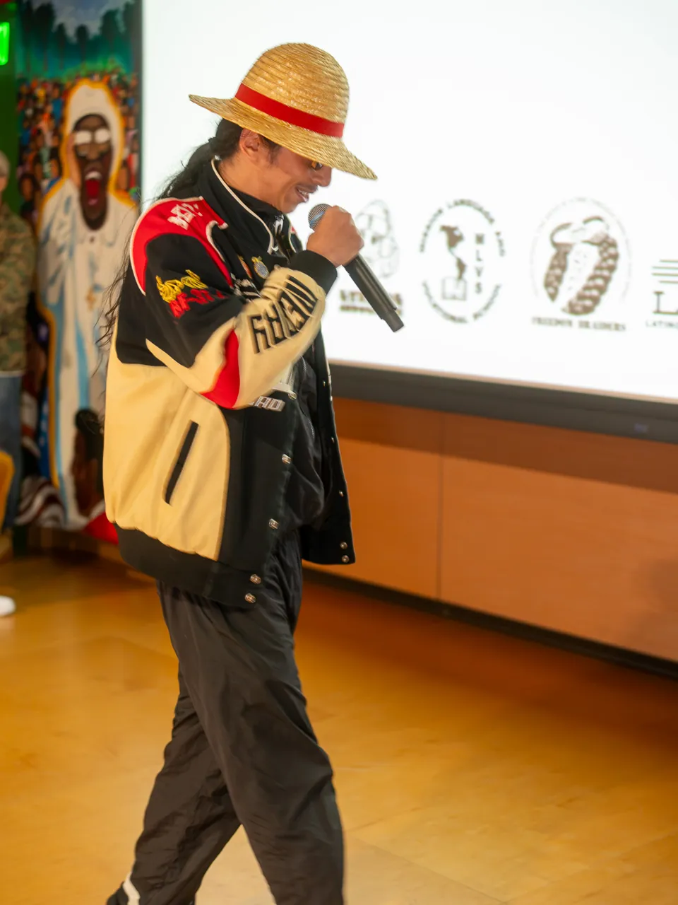 A performer in a straw hat and racing jacket delivers into a microphone beside the projection screen.