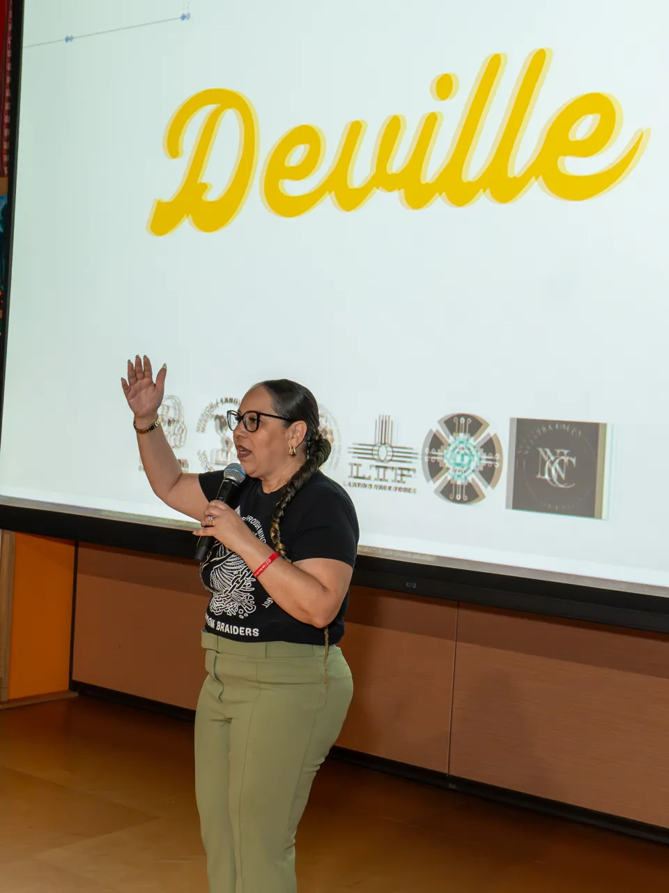 A speaker in a black shirt and olive trousers gestures with a raised hand beneath the Deville projection.