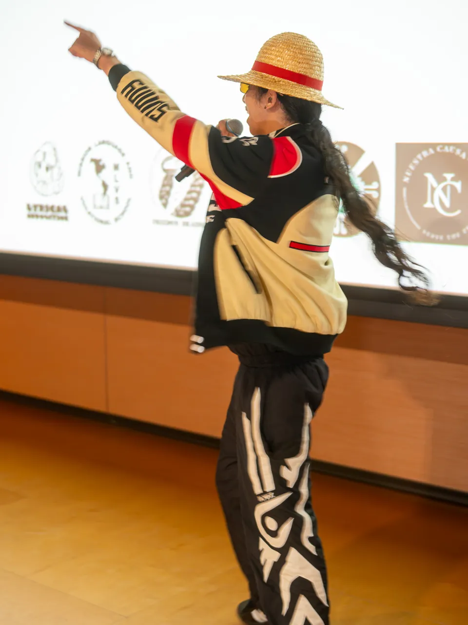 A performer in a straw hat raises an arm while holding a microphone beside the screen.