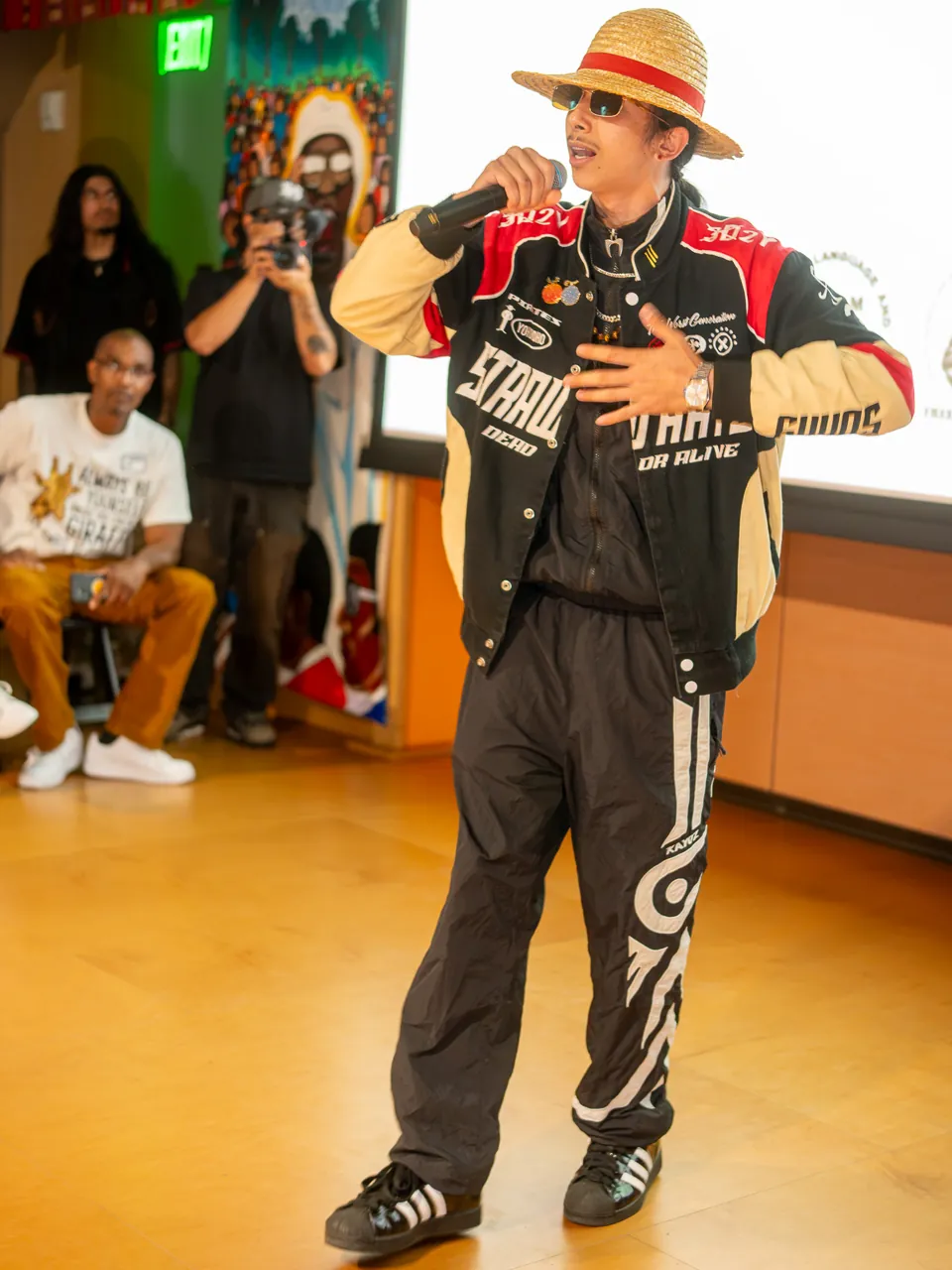 A performer in a straw hat and racing jacket delivers into a microphone beside the projection screen.
