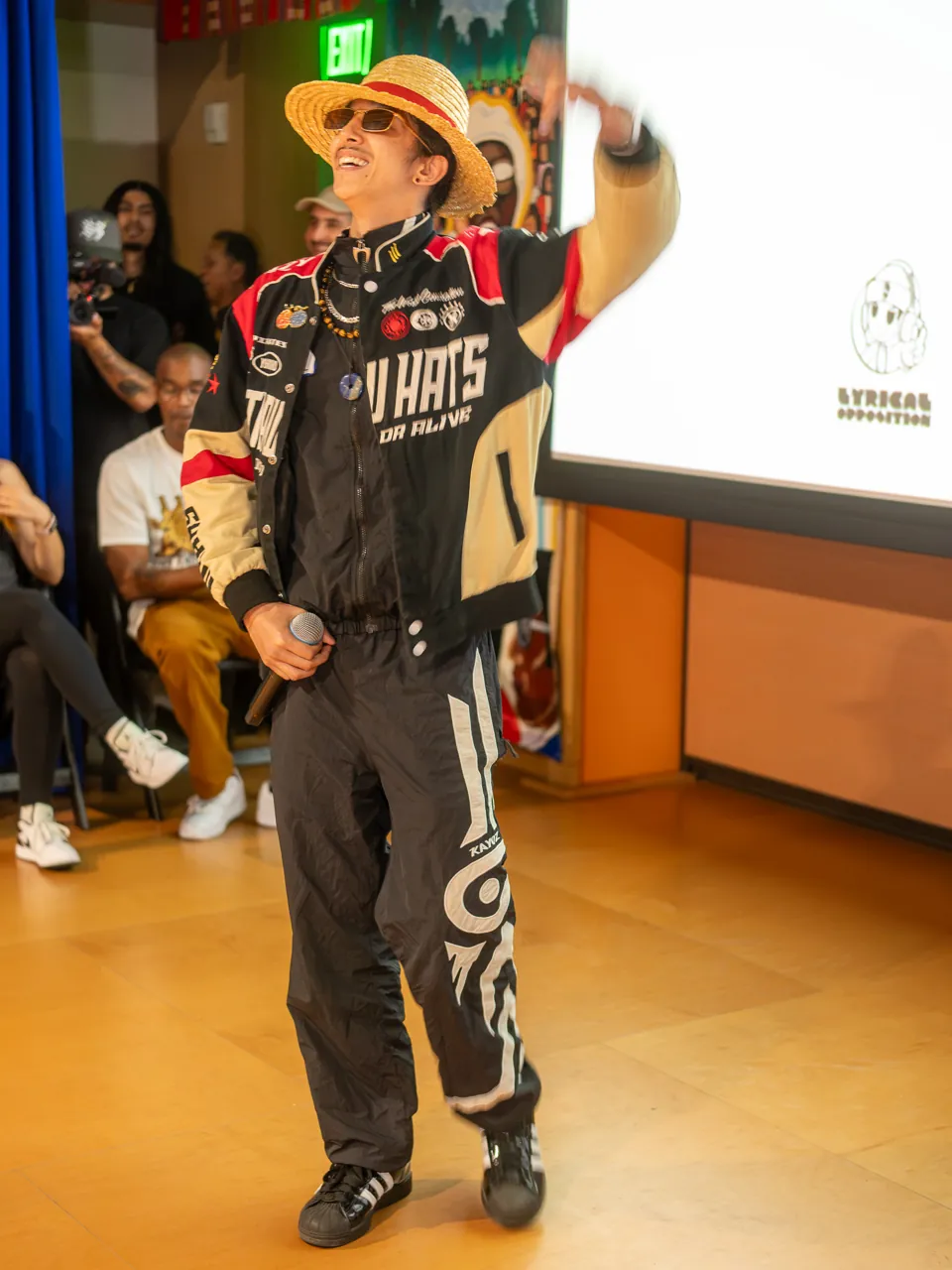 A performer in a straw hat raises an arm while holding a microphone beside the screen.